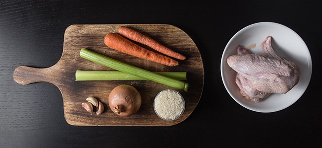 cutting board with 2 unpeeled carrots, 2 stalks of celery, 3 cloves of garlic, 1 onion, and a cup of uncooked rice. Next to it is a bowl of raw chicken.