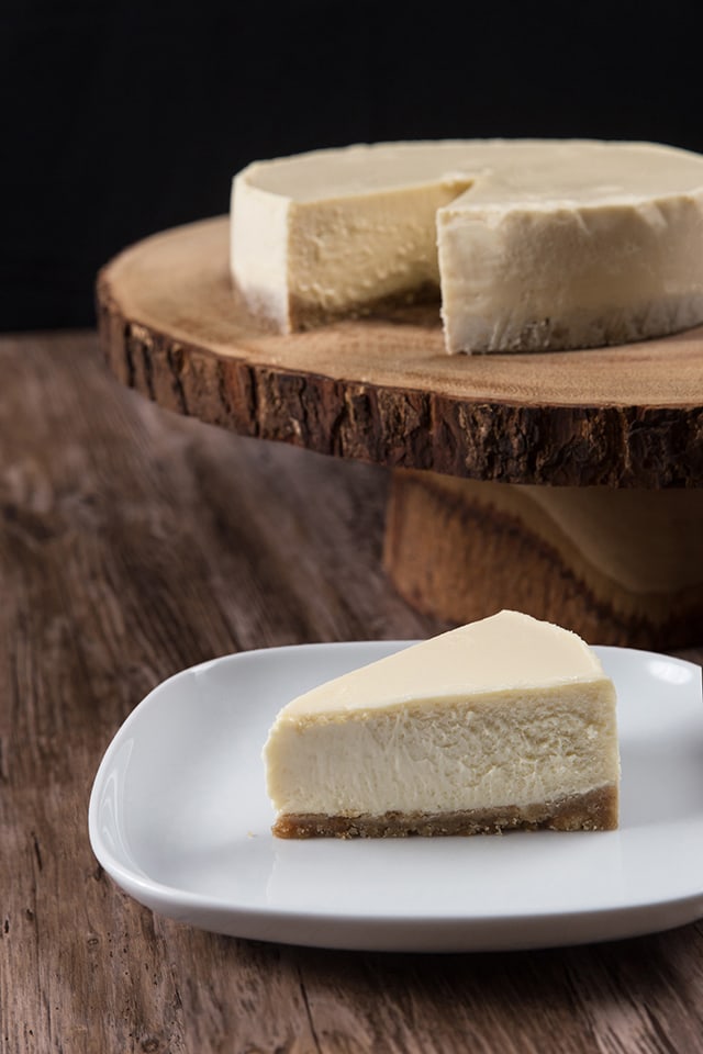 a slice of  New York Cheesecake made in a pressure cooker on a white plate with rest of cheesecake in the background on a tree trunk syle cake stand