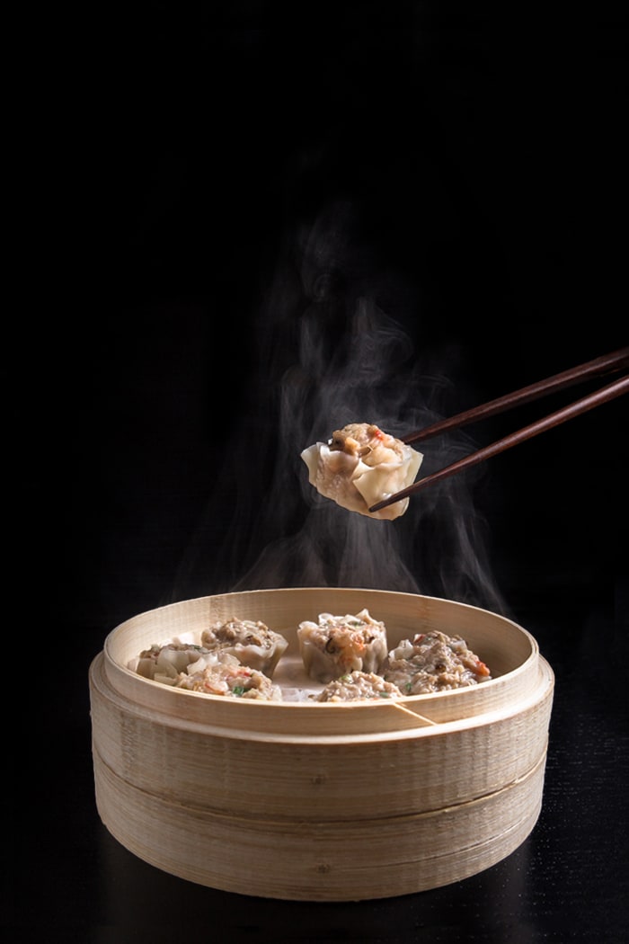 Shumai (Shrimp &amp; Pork Dumplings) in a steamer basket. One dumpling being held with chopsticks