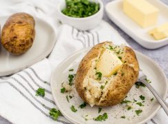 Instant pot baked potatoes with a pat of butter and fresh herbs sprinkled on top on a white plate with a butter dish in the background