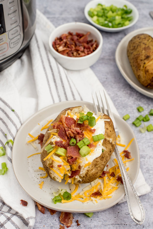 instant pot baked potatoes topped with scallions, shredded cheese and crumbled bacon on a white plate