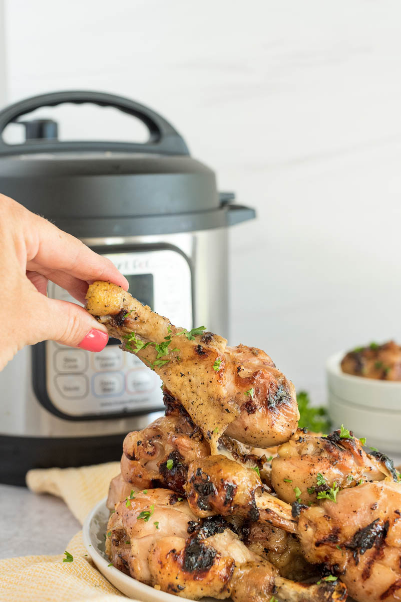 A hand grabbing a drumstick from a plate of honey Sriracha drumsticks placed in front of an Instant Pot.