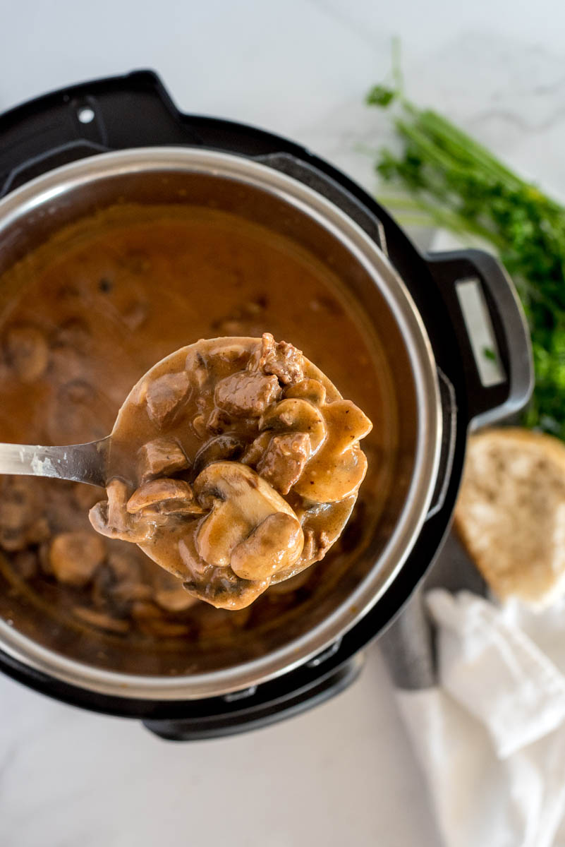 A ladle full of beef stroganoff with mushrooms visible suspended over an Instant Pot