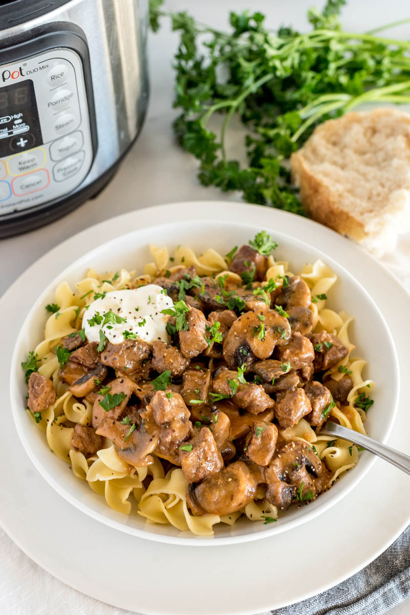 A 45 degree shot of pressure cooker beef stroganoff with parsley, a roll, and an instant Pot in the background