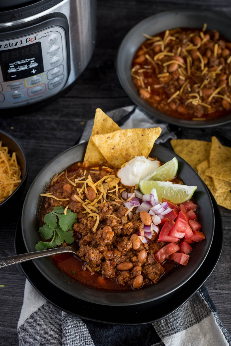 Picture of Instant Pot chili recipe with diced tomatoes, red onions, lime wedges, sour cream, cilantro, and tortilla chips in a bowl, with another bowl in the background and an Instant Pot..