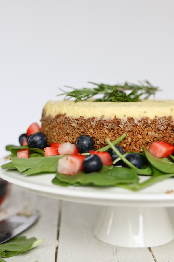 cake plate holding a savory cheesecake made with blue cheese, surrounded by a spinach salad with blueberries and strawberries