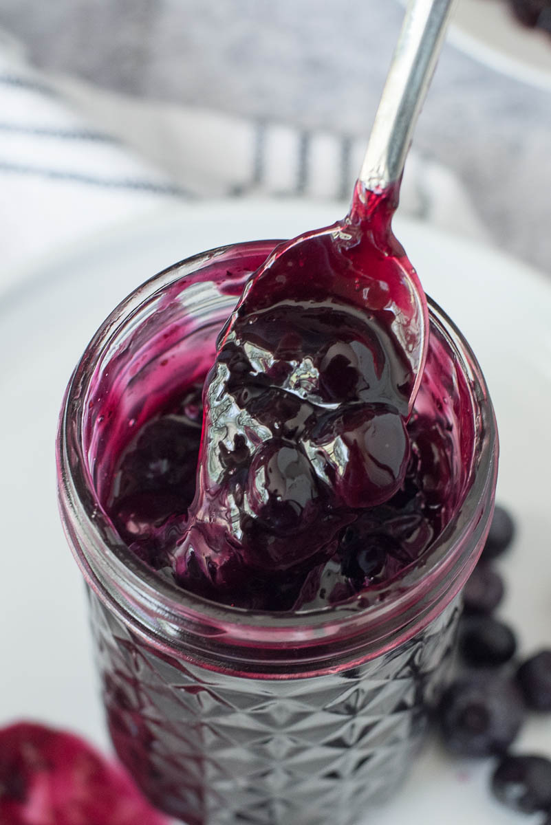 a textured mason jar filled with blueberry compote with extra blueberries on a white plate with a stack of pancakes topped with blueberry sauce in the background