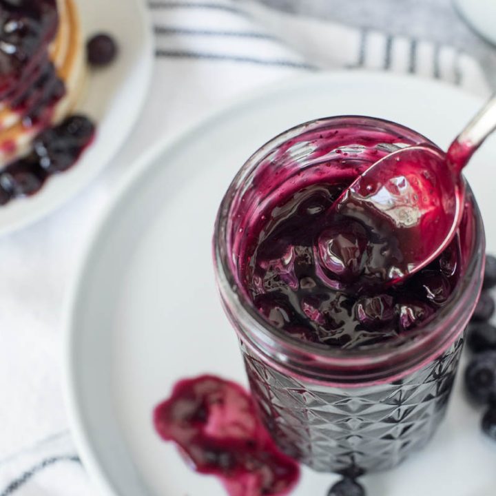 Overhead picture with textured mason jar filled with blueberry compote with extra blueberries on a white plate with a stack of pancakes topped with blueberry sauce in the background