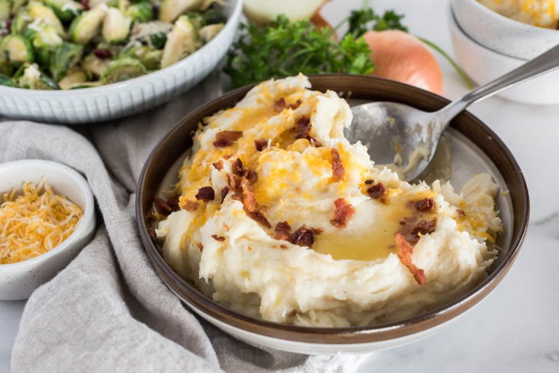 A 45 degree shot of a silver spoon dipping into a bowl of caramelized onion mashed potatoes, topped with shredded cheese and bacon