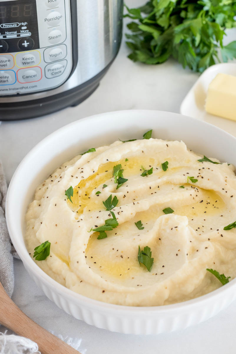 a 45 degree shot of mashed cauliflower in a white shallow bowl, topped with melted butter, parsley, and ground pepper, with an Instant Pot in the background