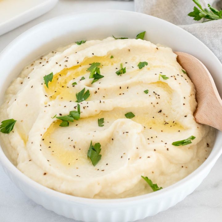 A 45 degree shot of a shallow white bowl filled with a swirled batch of cauliflower mash garnished with butter and parsley.