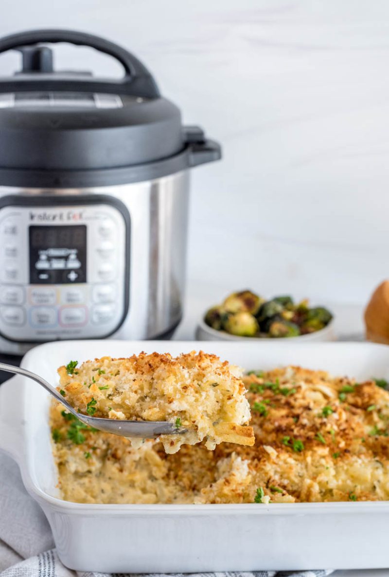 Pressure cooker hash brown casserole being scooped from a white serving dish, placed in front of an Instant Pot.