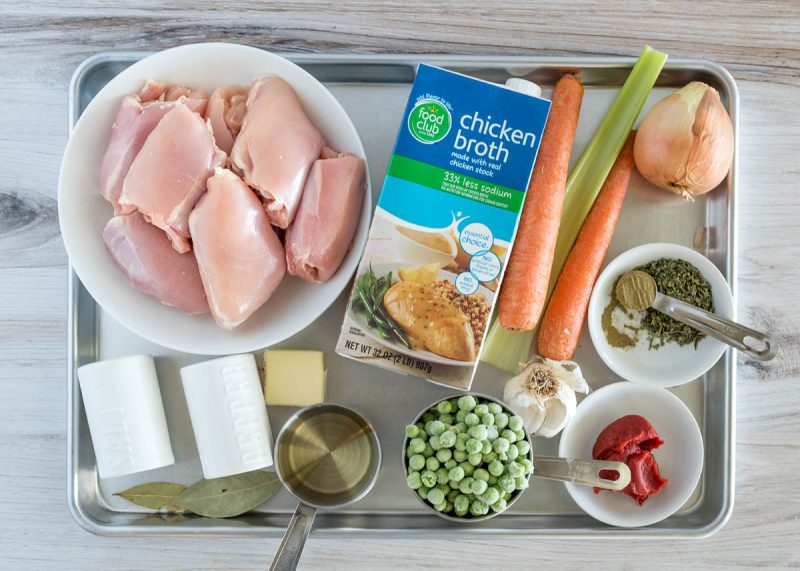 Overhead picture of ingredients for Instant Pot chicken and dumplings, including chicken thighs, chicken broth, carrots, celery, onion, seasonings, tomato paste, garlic, frozen peas, oil, butter, and bay leaves.