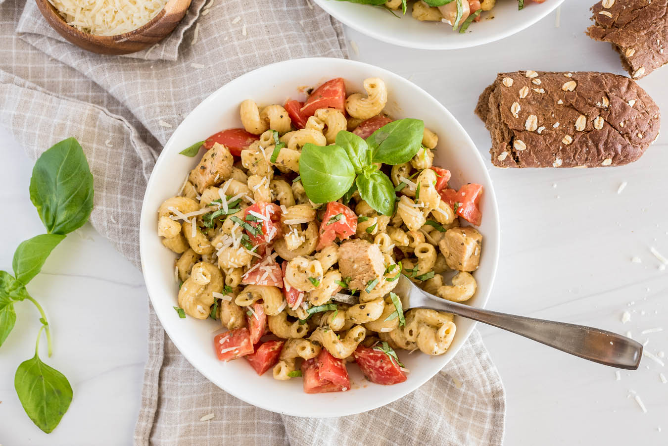 Over head shot of a white bowl with Instant Pot chicken pesto pasta topped with fresh basil and parmesan cheese, with bread and more basil in the background.