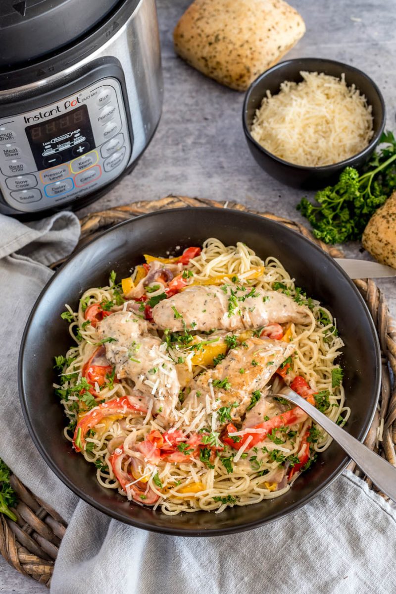 an overhead shot of a black bowl of chicken scampi pasta on a wooden charger with an instant pot and rolls in the background