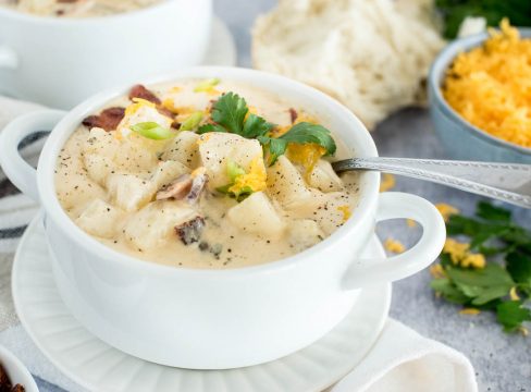 A 45 degree shot of Instant Pot potato soup, made with chunky potatoes, cheddar cheese, and bacon, served in a white soup bowl with a silver spoon tucked in the top right side of the bowl with cheese and bread in the background