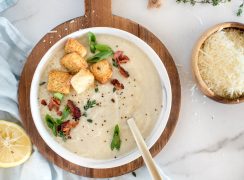 An overhead shot of a white bowl of cauliflower soup topped with croutons, diced green onions, bacon, and extra salt and pepper