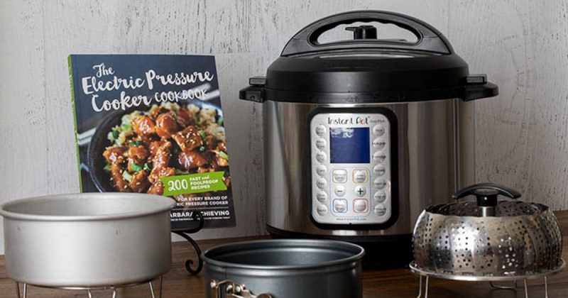 The Electric Pressure Cooker Cookbook on a cookbook stand, next to an Instant Pot Duo Plus, a cake pan, springform pan, and steamer basket—all essentials for using your new Instant Pot or electric pressure cooker