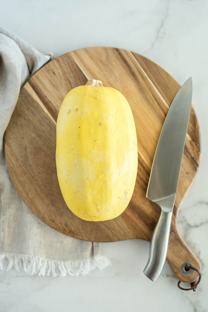 an overhead shot of a yellow oblong spaghetti squash on a wooden cutting board with a silver knife and a neutral linen towel