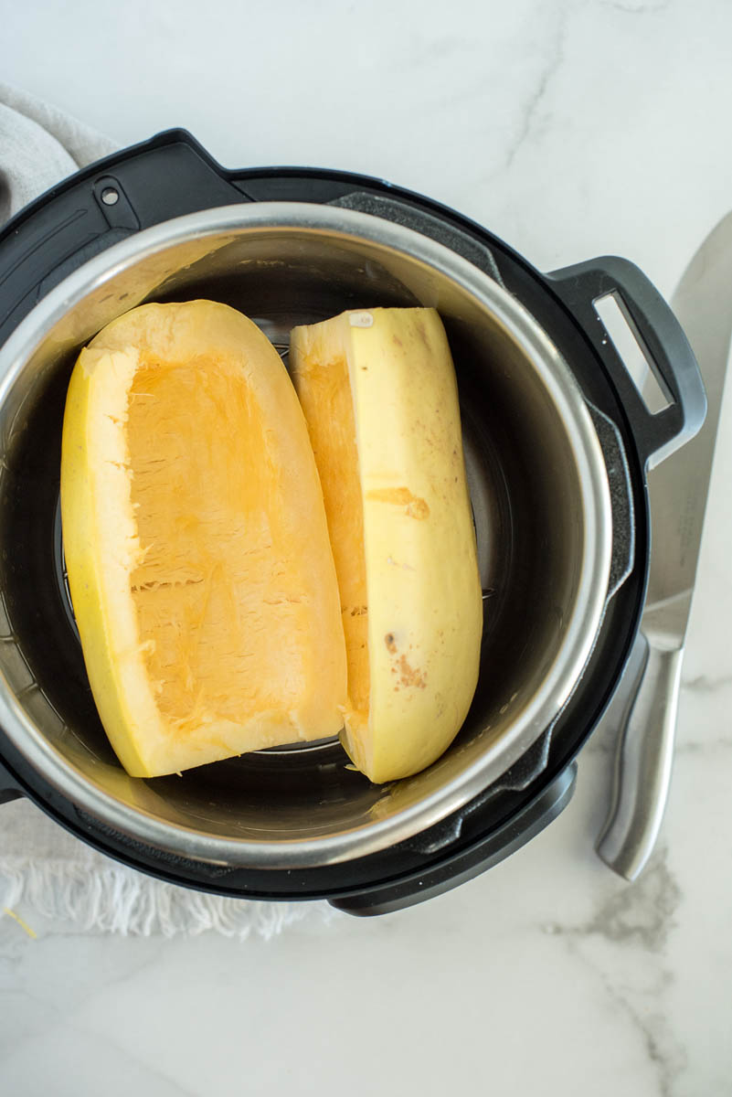 an overhead shot looking into an Instant Pot filed with the two cleaned spaghetti squash halves, with the knife and the linen towel in the background