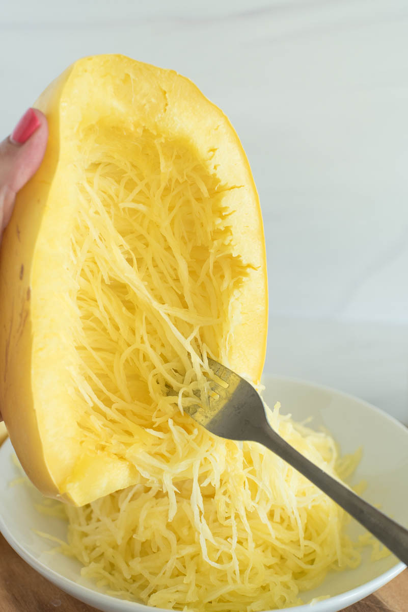 a side shot showing a hand holding the cooked spaghetti squash over a white bowl, with a fork scraping the "spaghetti" strands from the squash into the bowl