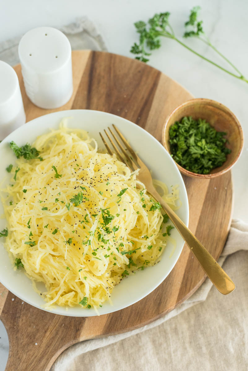 an overhead shot of a shallow bowl filled with spaghetti squash "noodles", along with a fork and a bowl of parsley garnish, with salt and pepper shakers in the background