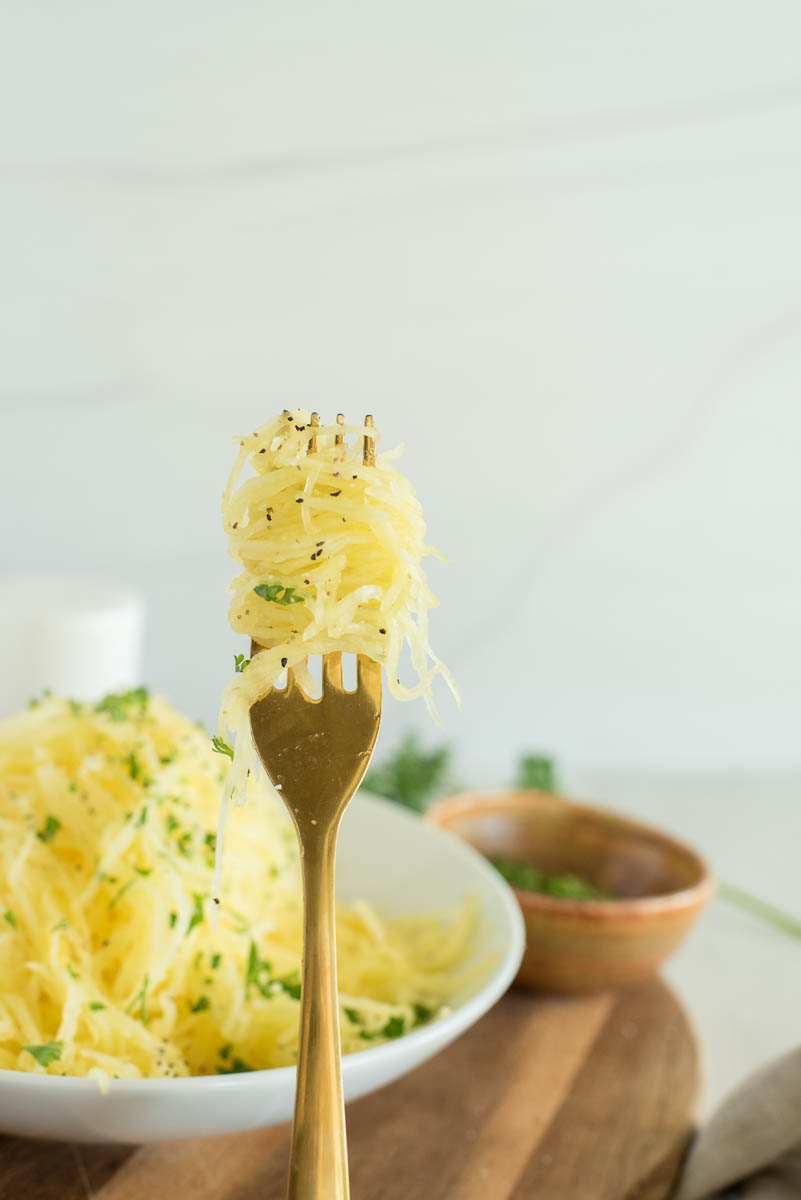 a straight-on shot of a fork with spaghetti squash noodles twirled around it, with a bowl of prepared spaghetti squash in the background
