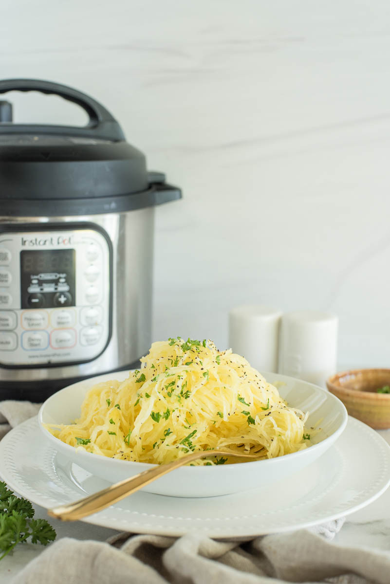 a vertical shot of a white bowl of spaghetti squash, sitting on a rimmed white plate with a fork in the front and an Instant Pot and salt and pepper shakers in the background