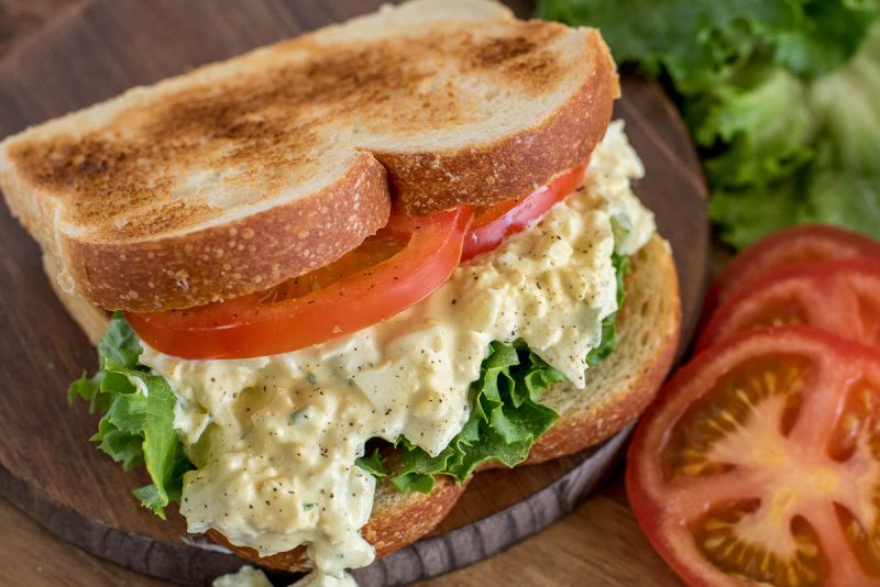 Close up picture of pressure cooker egg salad sandwich made with toasted bread, sliced tomato, and lettuce. Placed on a wooden cutting board.