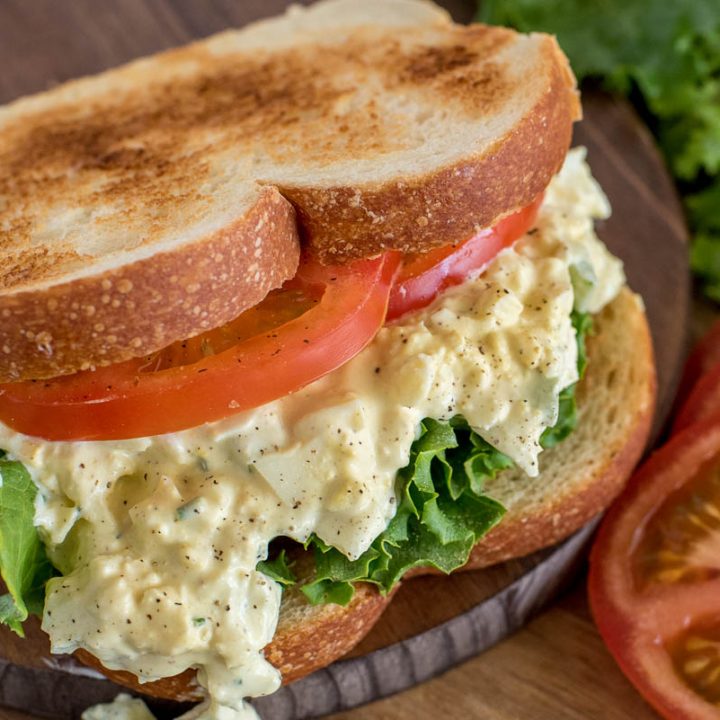 Close up picture of pressure cooker egg salad sandwich made with toasted bread, sliced tomato, and lettuce. Placed on a wooden cutting board.