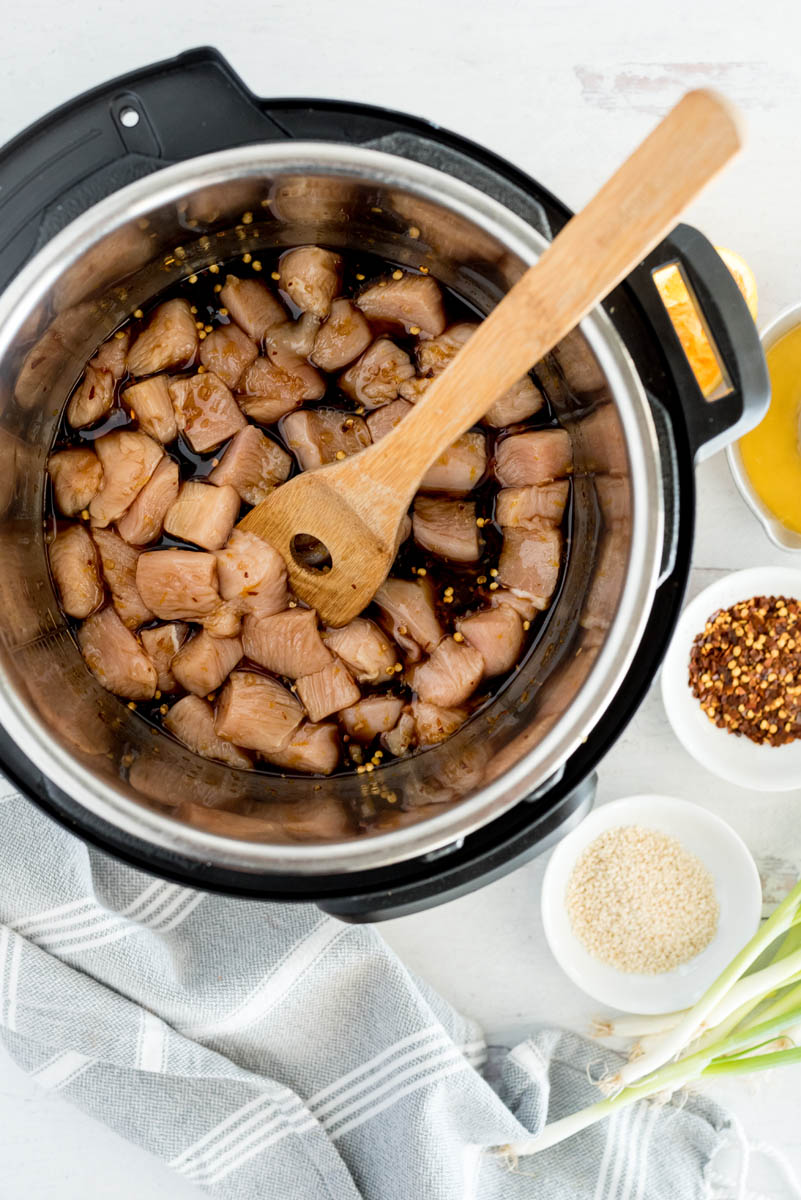 an overhead shot looking into an Instant Pot filled with diced chicken, teriyaki sauce, and sesame seeds, with a long wooden spoon in the top right of the image