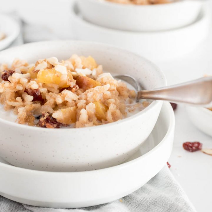a white bowl of Instant Pot Apple Risotto with Cherries and diced apples, with a silver spoon sticking out the side and topped with additional milk