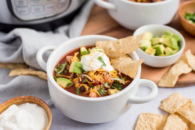 Close up picture of chicken enchilada soup topped with cheese, avocado, sour cream and chips. Placed in front of an Instant Pot.
