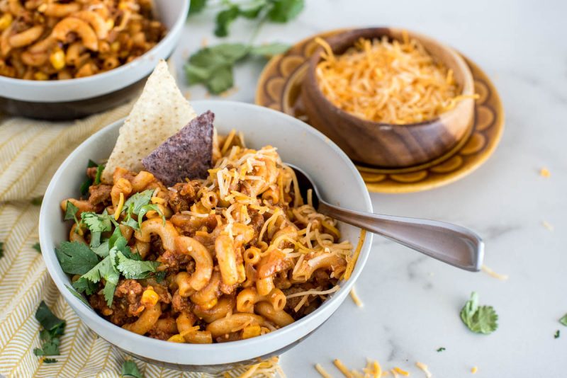 Close-up shot of Instant Pot Chili Pasta, served in a white bowl and garnished with cilantro, shredded cheese, and white and blue corn chips, with a bowl of shredded cheese in the background