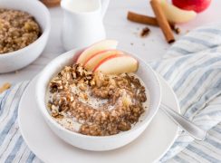 white bowl of apple-cinnamon flavored instant pot steel cut oats with apple slices, on top of a muted blue and white striped napkin with a glass of milk and a second bowl of steel cut oats in the background