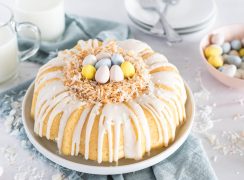 Instant Pot Coconut Bundt Cake, drizzled with white glaze and decorated with toasted coconut to look like a nest and Cadbury mini eggs on top, against a backdrop of a blue napkin and two classes of milk and a bowl of mini eggs in the background