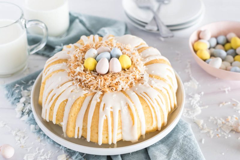 Instant Pot Coconut Bundt Cake, drizzled with white glaze and decorated with toasted coconut to look like a nest and Cadbury mini eggs on top, against a backdrop of a blue napkin and two classes of milk and a bowl of mini eggs in the background