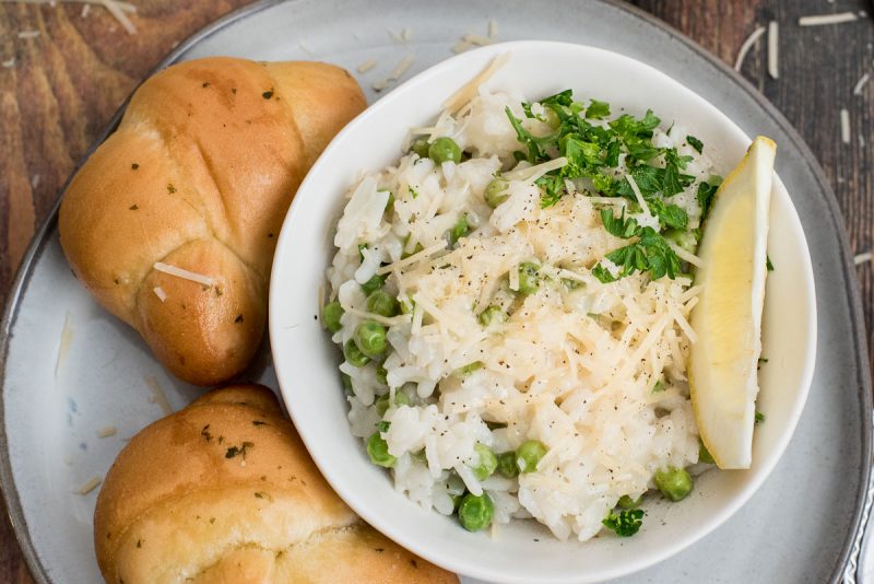 Close up overhead picture of lemon risotto made in an Instant Pot with peas, fresh parsley, parmesan, and a lemon wedge, next to some rolls.