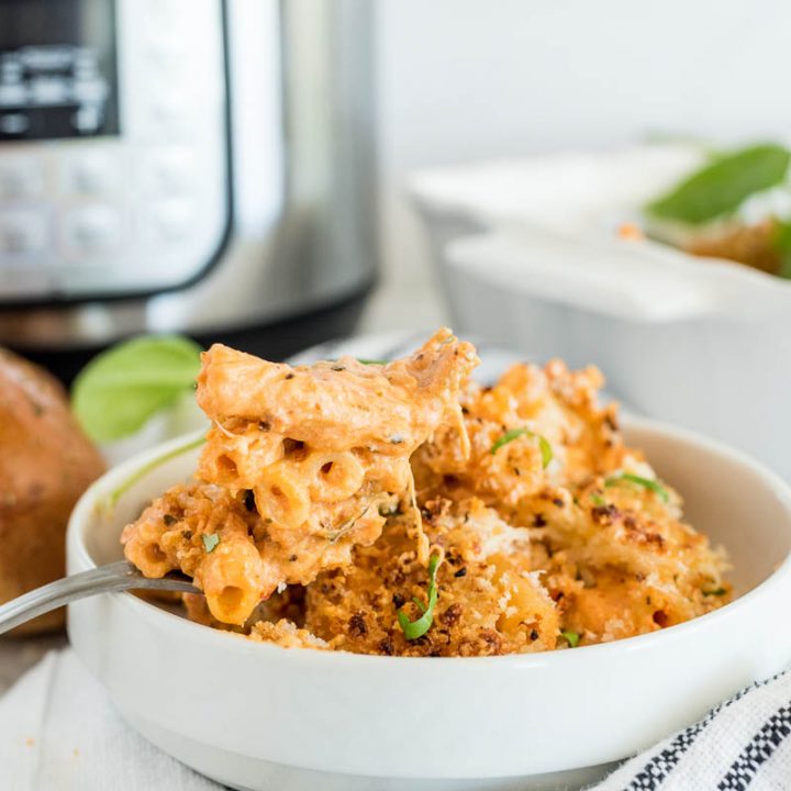 A horizontal shot of a small white bowl filled with cooked ziti and a spoon, sitting in front of an Instant Pot and several rolls