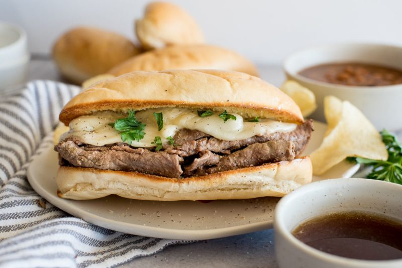 A close up of an Instant Pot French dip sandwich topped with cheese on a bun next to a bowl of au jus.