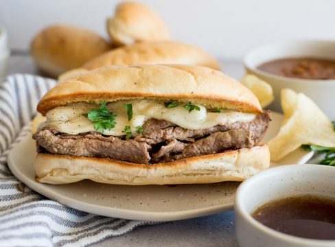 A close up of an Instant Pot French dip sandwich topped with cheese on a bun next to a bowl of au jus.