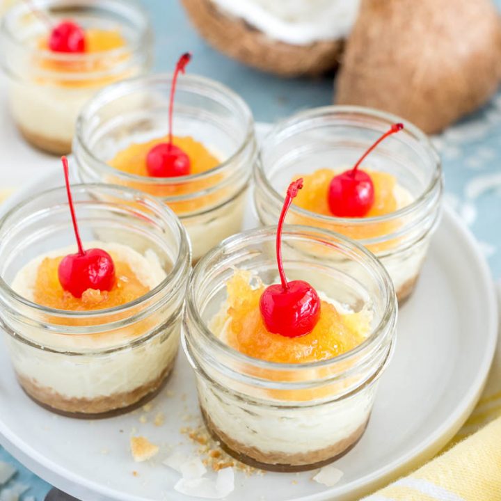 Four mini mason jars filled with mini tropical cheesecakes, topped with crushed pineapple topping and a maraschino cherry on top