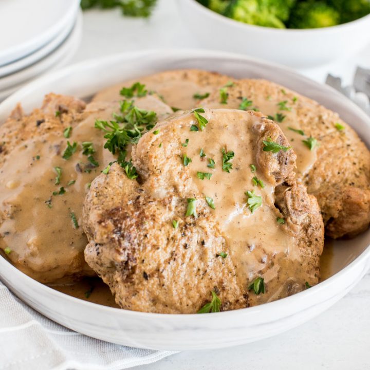 close up on a white bowl with four pork chops in mushroom gravy with steamed broccoli in the background