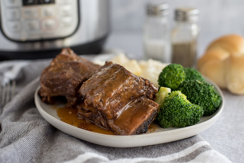 Bone-in pressure cooker Short Ribs plated with broccoli and mashed potatoes in front of an Instant Pot.