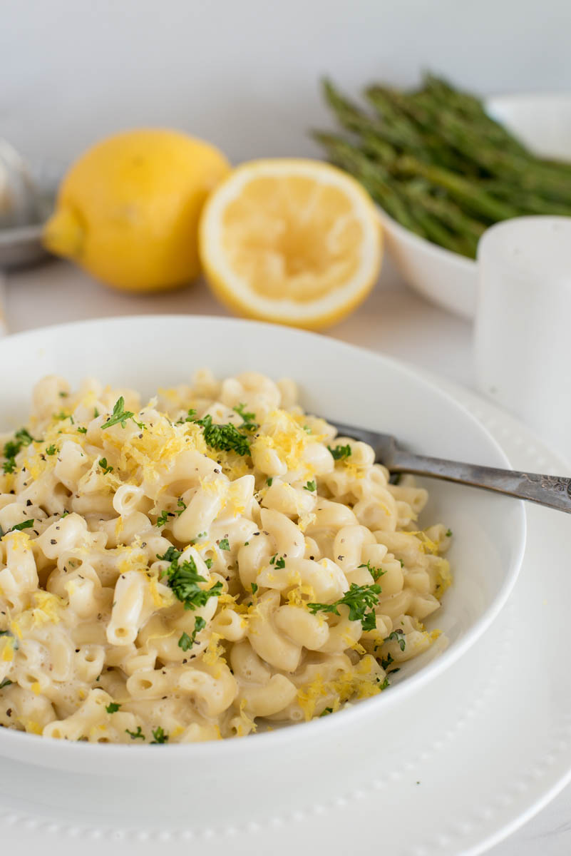a 45 degree vertical shot of a white bowl filled with creamy macaroni, garnished with lemon and parsley, with additional lemon and a bowl of asparagus in the background