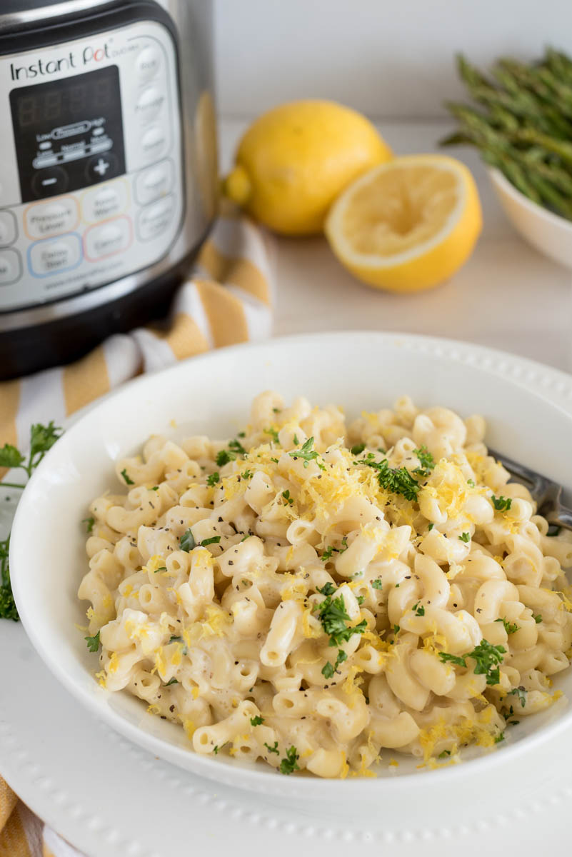 a 45 degree shot of a white bowl filled with Instant Pot lemon pasta, garnished with parsley, and sitting in front of an Instant Pot with juiced lemons and asparagus in the background