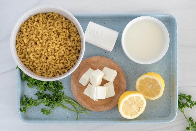 an overhead shot of the ingredients needed to make lemon pasta, including macaroni, cream cheese, salt, lemon, and evaporated milk on a muted teal baking sheet with green parsley for garnish