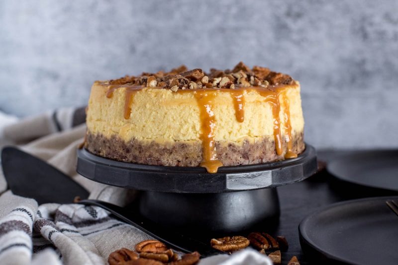 A side view of an Instant Pot New York cheesecake on a cake stand and topped with caramel, toffee, and pecans.