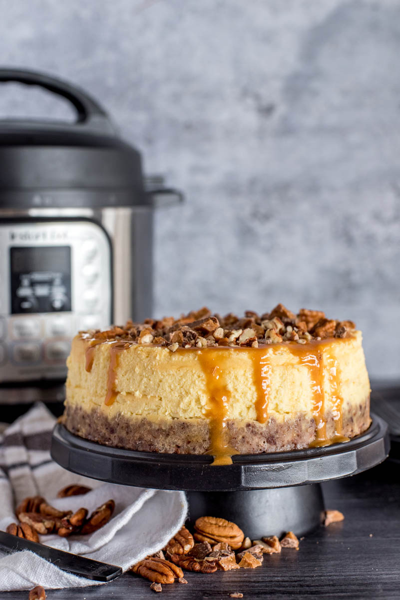 A New York cheesecake on a cake stand topped with Carmel, toffee, and pecans, and placed in front of an Instant Pot.