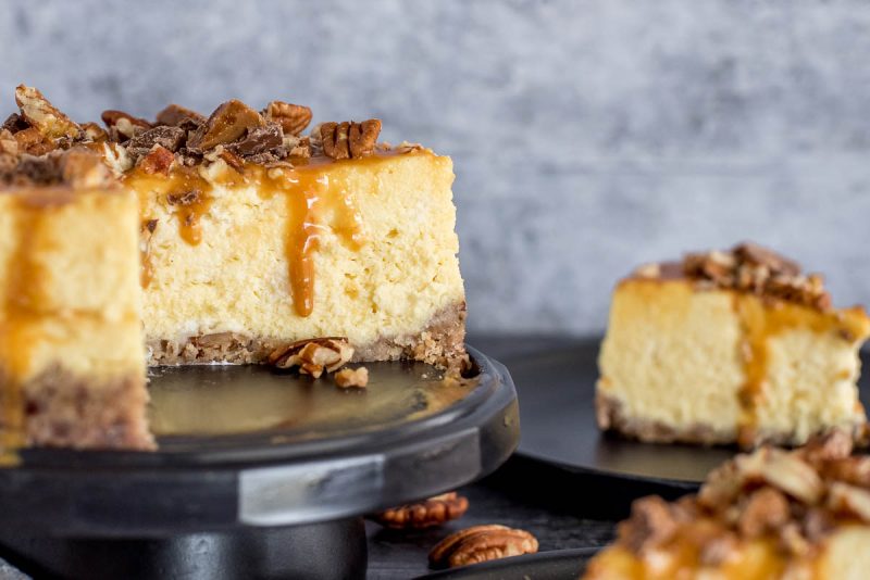 New York cheesecake on a cake stand topped with Carmel and pecans, with a slice in the background.